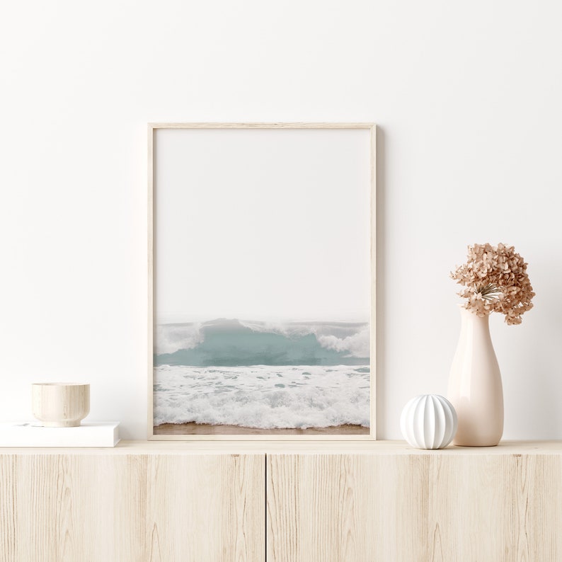 May include: A framed photograph of ocean waves crashing on a sandy beach. The photo is in a light wood frame and is hanging on a white wall. There is a vase with dried flowers and a small white decorative object on a wooden shelf below the photo.