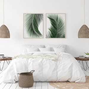 K&ouml;nnte beinhalten: Zwei gerahmte Drucke von gr&uuml;nen Palmbl&auml;ttern an einer wei&szlig;en Wand &uuml;ber einem wei&szlig;en Bett mit einer wei&szlig;en Bettdecke und einem braunen Weidenkorb.
