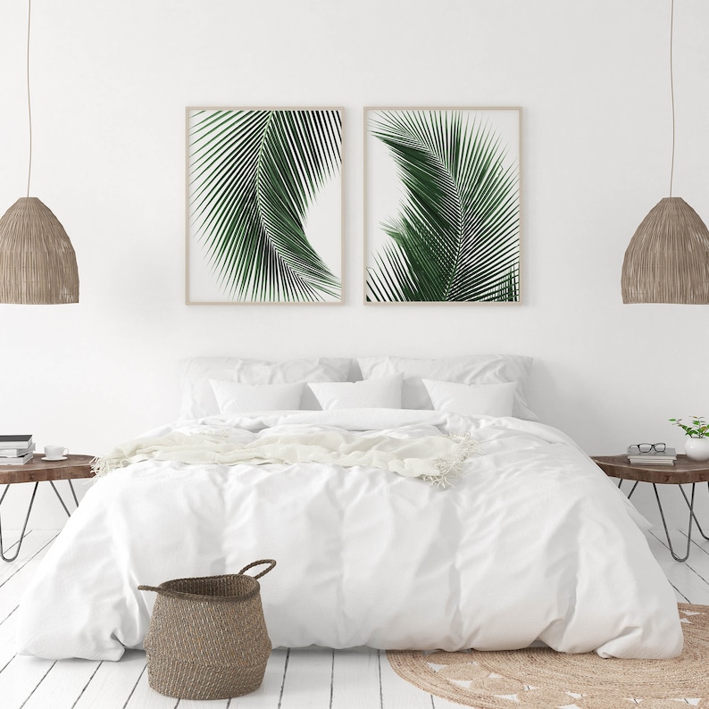K&ouml;nnte beinhalten: Zwei gerahmte Drucke von gr&uuml;nen Palmbl&auml;ttern an einer wei&szlig;en Wand &uuml;ber einem wei&szlig;en Bett mit einer wei&szlig;en Bettdecke und einem braunen Weidenkorb.