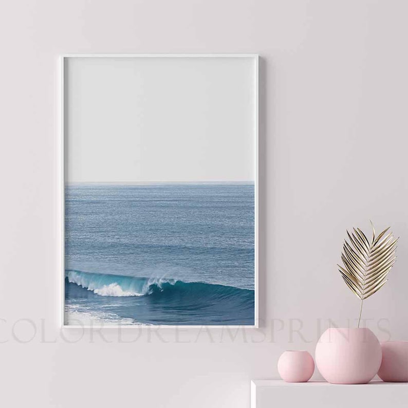 May include: A framed photograph of a blue ocean wave crashing against the shore. The wave is white and foamy, and the ocean is a deep blue. The sky is a light blue, and there is a white border around the photograph.