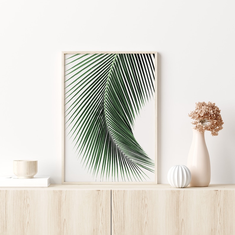 K&ouml;nnte beinhalten: Ein gerahmter Druck eines einzelnen gr&uuml;nen Palmblattes vor wei&szlig;em Hintergrund. Der Druck wird auf einem hellen Holzregal mit einer kleinen wei&szlig;en Schale und einer Vase mit getrockneten Blumen ausgestellt.