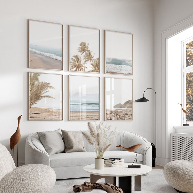 May include: A living room interior with a white couch and a white coffee table. Six framed prints of beach scenes with palm trees and ocean waves hang on the wall. A tall floor lamp and a small wooden bird sculpture are also visible.