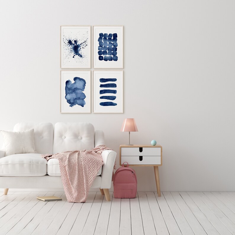 Puede incluir: Un sof&aacute; blanco con una manta de tiro rosa, una mesita de noche de madera con dos cajones, una l&aacute;mpara rosa y una mochila rosa. Cuatro pinturas abstractas de acuarela enmarcadas en tonos azules cuelgan de la pared blanca detr&aacute;s del sof&aacute;.