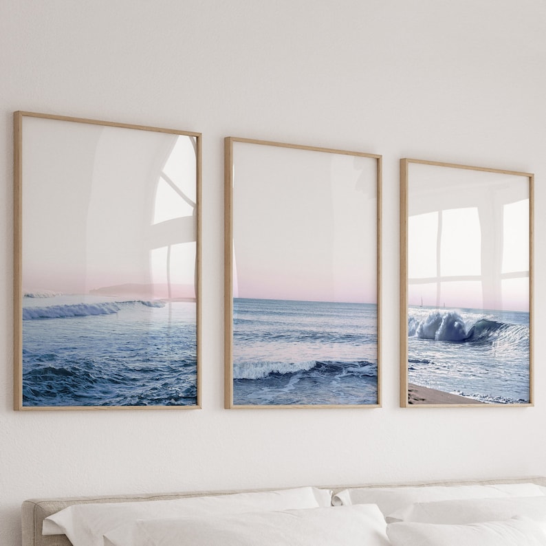 May include: Set of three framed art prints featuring ocean waves. The prints showcase a serene seascape with blue water, white waves, and a pink and white sky. The frames are a light brown color, adding a natural touch to the coastal theme.