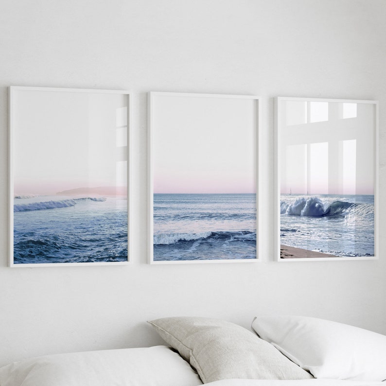 May include: Three framed art prints featuring ocean waves. The prints have white frames and depict the ocean with blue water and a pink and blue sky. The artwork is displayed above a bed with white sheets and pillows.