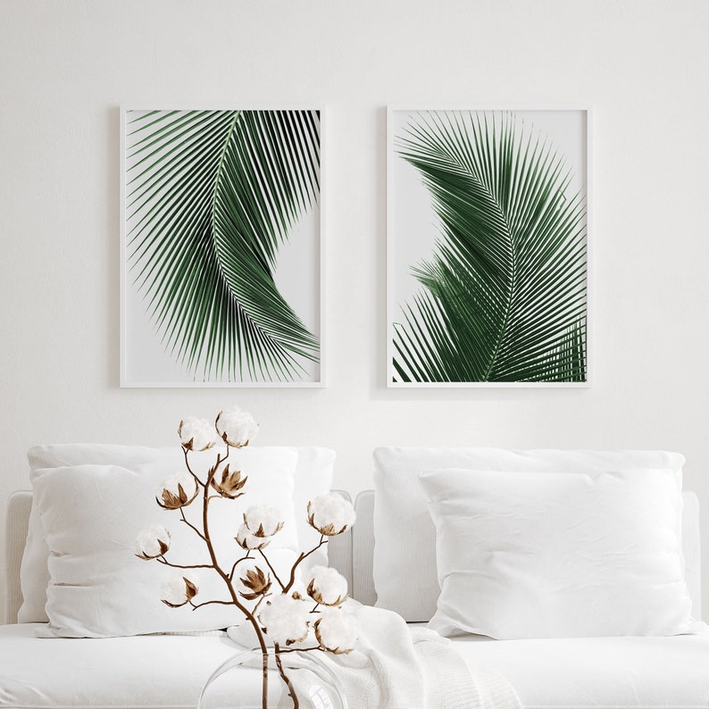 K&ouml;nnte beinhalten: Zwei gerahmte Drucke von gr&uuml;nen Palmbl&auml;ttern an einer wei&szlig;en Wand &uuml;ber einem wei&szlig;en Sofa mit wei&szlig;en Kissen und einem Baumwollzweig in einer Glasvase.