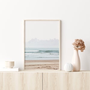 May include: A framed photograph of a beach scene with waves crashing on the shore. The photo is in a light wood frame and is hanging on a white wall.