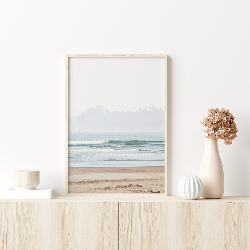 May include: A framed photograph of a beach scene with waves crashing on the shore. The photo is in a light wood frame and is hanging on a white wall.