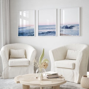 May include: Three framed ocean wave prints in white frames hang above two cream-colored armchairs. The prints depict the ocean with blue waves and a pink and blue sky. A round wooden coffee table and a vase of dried flowers are in front of the chairs.