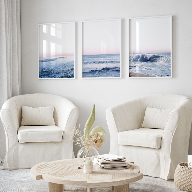 May include: Three framed ocean wave prints in white frames hang above two cream-colored armchairs. The prints depict the ocean with blue waves and a pink and blue sky. A round wooden coffee table and a vase of dried flowers are in front of the chairs.