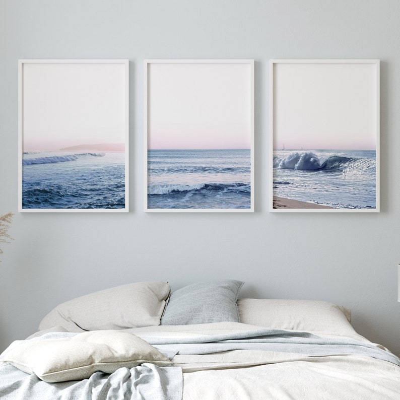 May include: Three framed art prints featuring ocean waves. The prints display a serene seascape with shades of blue and pink, capturing the movement of the water and the sky. The frames are white, and the overall aesthetic is calming.