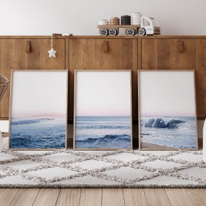May include: Three framed art prints featuring ocean wave photographs. The prints have light wood frames and depict various stages of waves in shades of blue and white, with a soft pink and blue sky. The prints are displayed on a wooden cabinet.