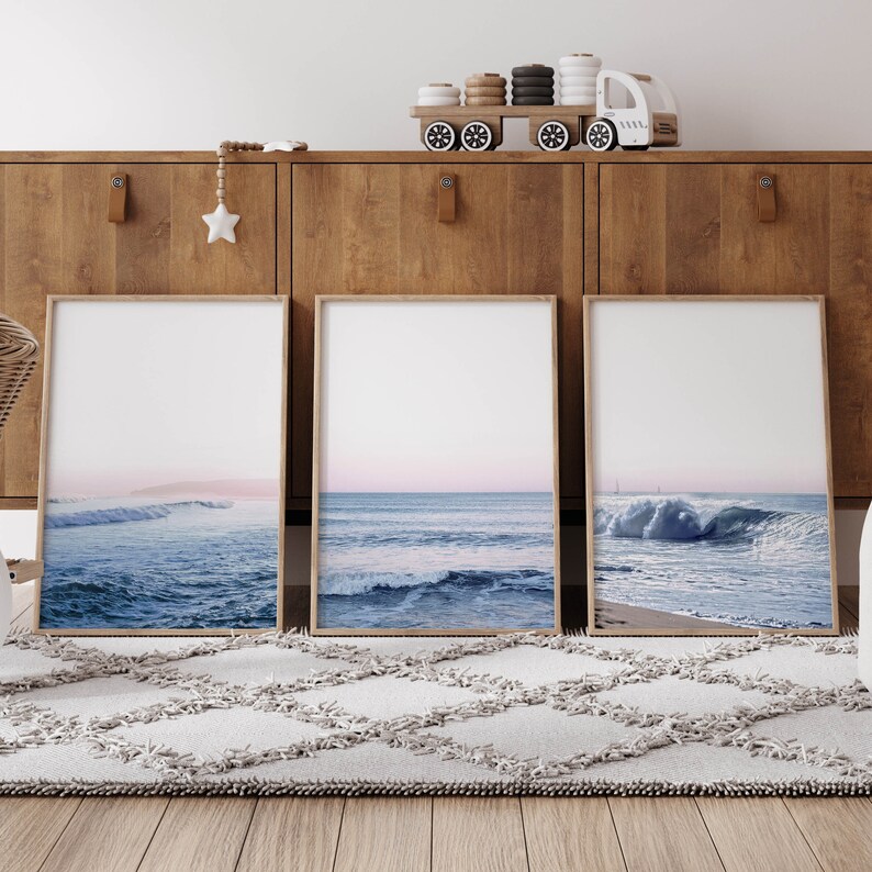 May include: Three framed art prints featuring ocean wave photographs. The prints have light wood frames and depict various stages of waves in shades of blue and white, with a soft pink and blue sky. The prints are displayed on a wooden cabinet.