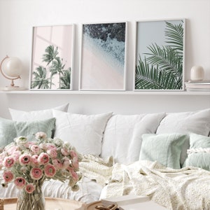Puede incluir: Tres impresiones enmarcadas con temas tropicales. La impresi&oacute;n de la izquierda presenta palmeras sobre un fondo rosa. La impresi&oacute;n del centro muestra una escena de playa con agua azul y arena blanca. La impresi&oacute;n de la derecha muestra hojas de palmera verdes sobre un fondo azul claro.