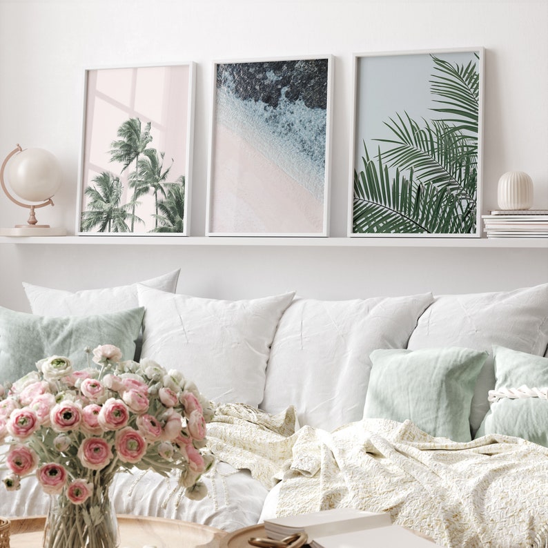 Puede incluir: Tres impresiones enmarcadas con temas tropicales. La impresi&oacute;n de la izquierda presenta palmeras sobre un fondo rosa. La impresi&oacute;n del centro muestra una escena de playa con agua azul y arena blanca. La impresi&oacute;n de la derecha muestra hojas de palmera verdes sobre un fondo azul claro.