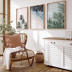 Puede incluir: Una silla de rat&aacute;n con marco de madera clara, una manta de tiro marr&oacute;n y una almohada de color tostado. La silla est&aacute; frente a una pared blanca con tres impresiones enmarcadas de plantas tropicales y un dise&ntilde;o abstracto azul claro. Un armario blanco con una tapa de madera y dos cajones est&aacute; en el lado derecho de la imagen.
