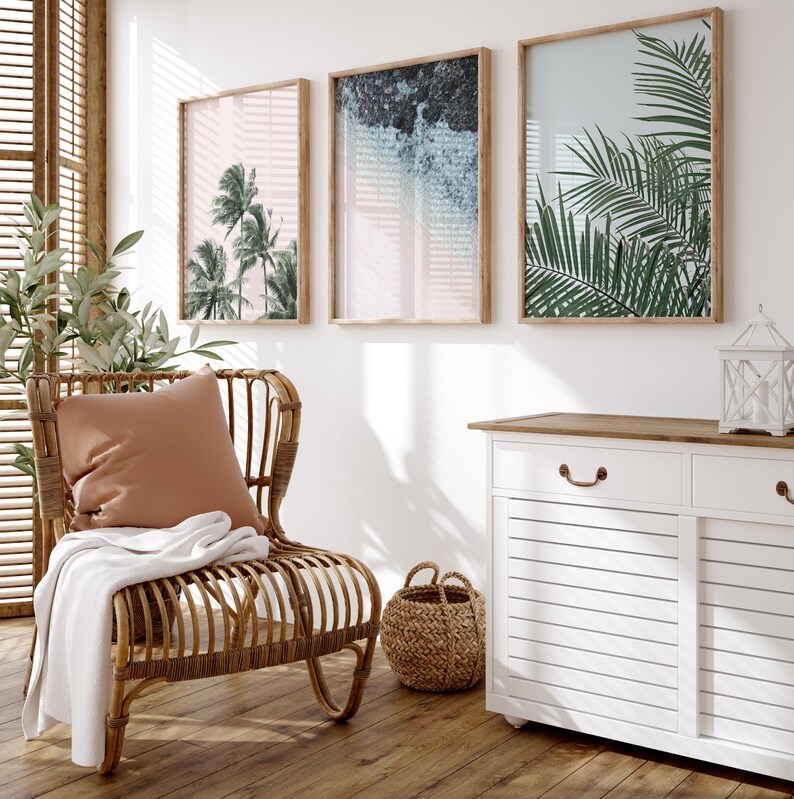 Puede incluir: Una silla de rat&aacute;n con marco de madera clara, una manta de tiro marr&oacute;n y una almohada de color tostado. La silla est&aacute; frente a una pared blanca con tres impresiones enmarcadas de plantas tropicales y un dise&ntilde;o abstracto azul claro. Un armario blanco con una tapa de madera y dos cajones est&aacute; en el lado derecho de la imagen.