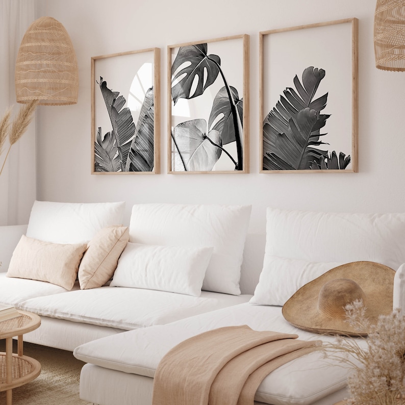 Pode incluir: Tr&ecirc;s impress&otilde;es em preto e branco de folhas tropicais emolduradas penduradas em uma parede branca acima de um sof&aacute; branco com almofadas e um cobertor bege.