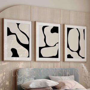 May include: Three framed abstract art prints. Each print features a beige background with black organic shapes. The frames are a light wood color, and the prints are displayed above a bed with a blue headboard and pillows.