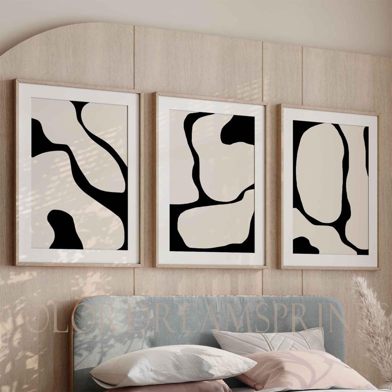 May include: Three framed abstract art prints. Each print features a beige background with black organic shapes. The frames are a light wood color, and the prints are displayed above a bed with a blue headboard and pillows.