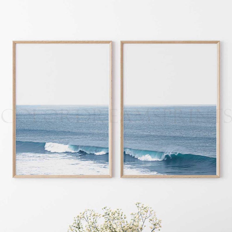 May include: Two framed prints of a blue ocean with white waves crashing on the shore. The prints are in wooden frames and are hung on a white wall.