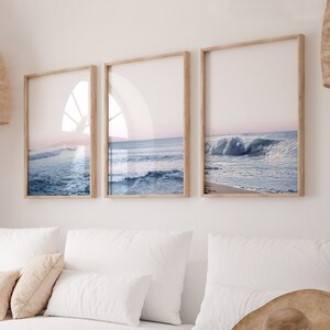 May include: Three framed prints of ocean waves. The prints have light wood frames and feature a serene seascape with blue water and a pink and blue sky. The artwork is displayed on a white wall.