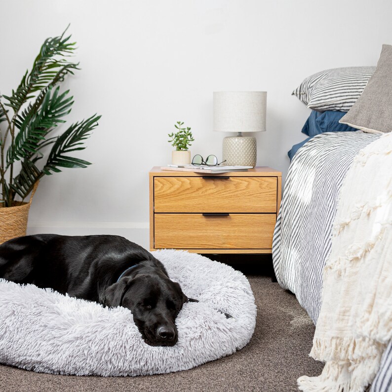 May include: A black dog sleeping on a gray fluffy dog bed in a bedroom with a wooden nightstand, a lamp, and a bed with striped bedding.