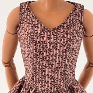 Może przedstawiać: Sukienka bez rękaw&oacute;w z dekoltem w serek z r&oacute;żowo-brązowym wzorem kwiatowym. Sukienka ma dopasowany gorset i rozkloszowaną sp&oacute;dnicę. Tkanina ma teksturowany wygląd, a kwiatowy wz&oacute;r powtarza się na całej odzieży. Sukienka jest przeznaczona dla lalki.