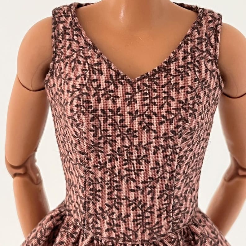 Może przedstawiać: Sukienka bez rękaw&oacute;w z dekoltem w serek z r&oacute;żowo-brązowym wzorem kwiatowym. Sukienka ma dopasowany gorset i rozkloszowaną sp&oacute;dnicę. Tkanina ma teksturowany wygląd, a kwiatowy wz&oacute;r powtarza się na całej odzieży. Sukienka jest przeznaczona dla lalki.