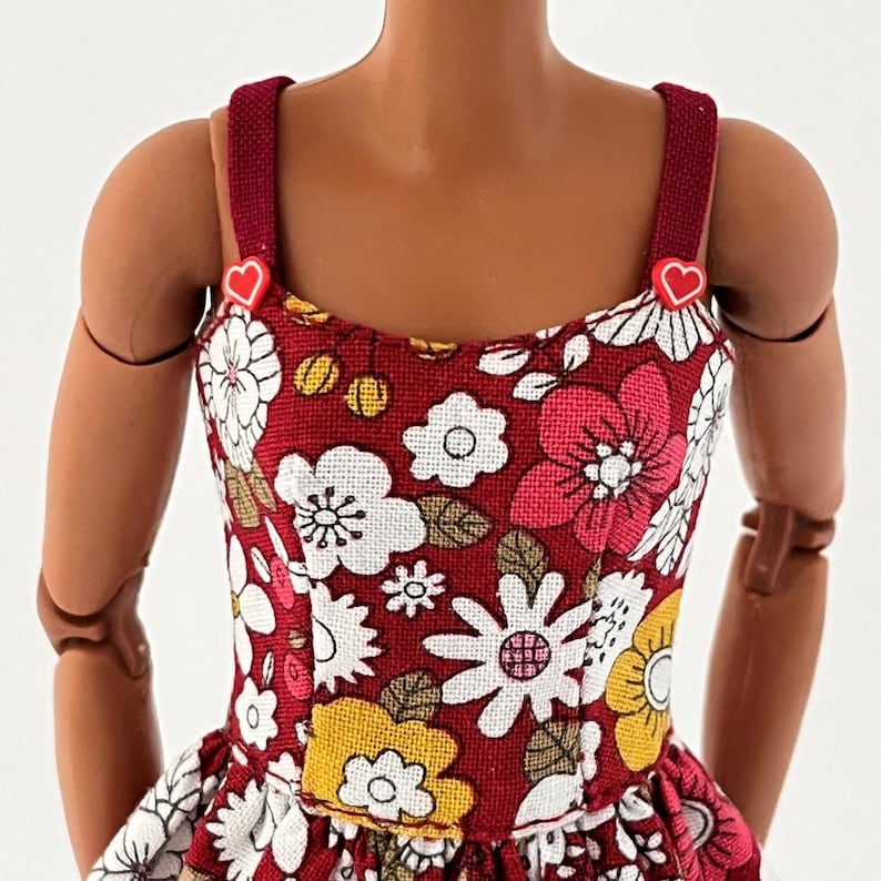 Może przedstawiać: Sukienka dla lalki z kwiatowym wzorem w kolorze czerwonym, białym, r&oacute;żowym i ż&oacute;łtym. Sukienka ma okrągły dekolt i czerwone ramiączka z akcentami w kształcie serca. Tkanina wydaje się być tkana.