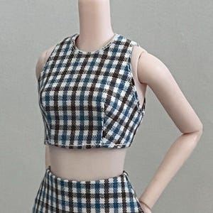 Puede incluir: Un conjunto de dos piezas en miniatura que consta de un top corto y una falda. La tela tiene un diseño a cuadros azul, blanco y marrón. El top corto tiene un cuello redondo y la falda es visible en la parte inferior de la imagen.