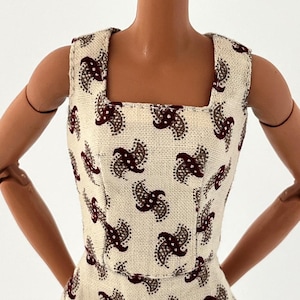 Może przedstawiać: Sukienka bez rękaw&oacute;w z kwadratowym dekoltem. Sukienka ma kremową tkaninę z powtarzającym się wzorem paisley w kolorze ciemnoczerwonym i szarym. Sukienka jest dopasowana i wydaje się być wykonana z teksturowanego materiału.