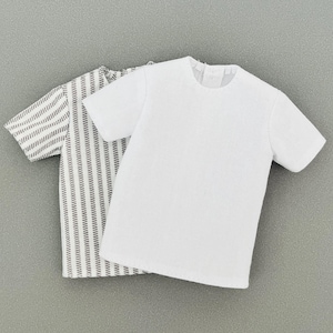 Könnte beinhalten: Zwei kurzärmelige Hemden. Eines ist weiß, das andere ist mit schwarz-weißen vertikalen Linien gestreift. Die Hemden sind aus einem weichen, wahrscheinlich Baumwollmaterial. Die Hemden liegen auf einer grauen Oberfläche.