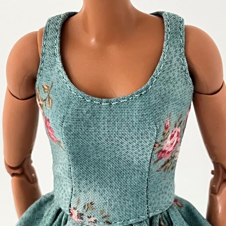 Może przedstawiać: Turkusowa sukienka dla lalek z kwiatowym wzorem przedstawiającym r&oacute;żowe r&oacute;że i zielone liście. Sukienka ma okrągły dekolt i jest obszyta białymi szwami. Tkanina ma teksturowany wygląd.