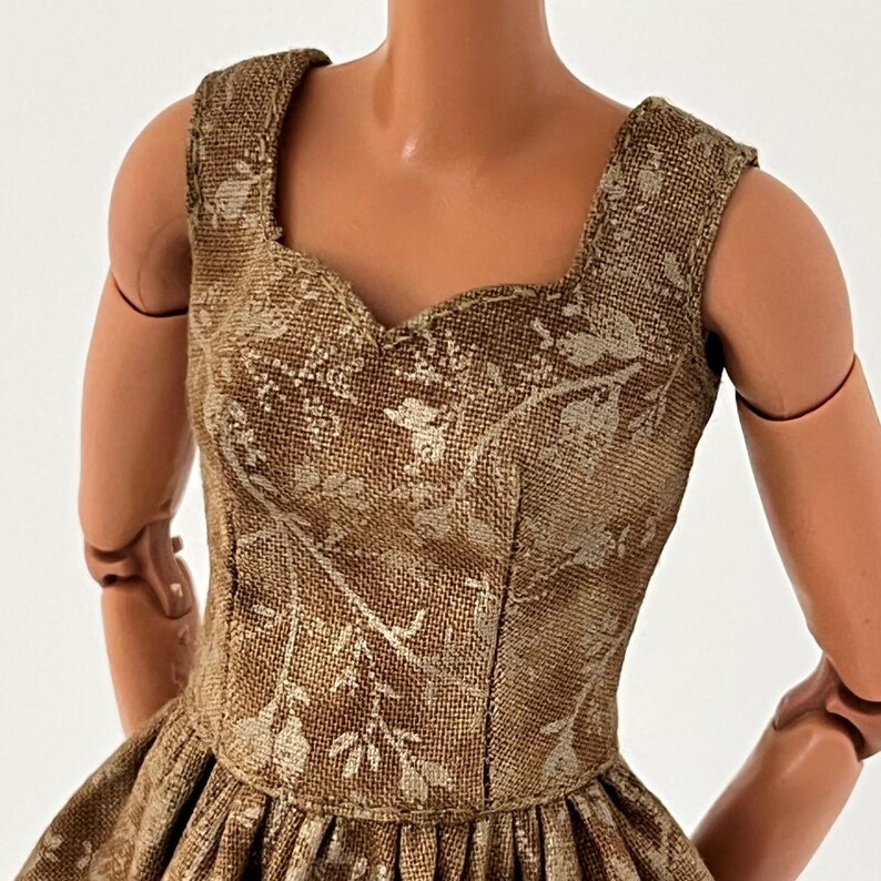 Może przedstawiać: Sukienka dla lalek we wz&oacute;r kwiatowy w kolorze beżowym i kremowym. Sukienka ma dekolt w kształcie serca i pełną sp&oacute;dnicę. Tkanina ma teksturowany wygląd, a sukienka jest bez rękaw&oacute;w. Ramiona lalki są widoczne po obu stronach sukienki.