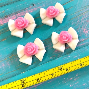 May include: Four white bow-shaped embellishments, each with a pink rose in the center. The bows are glossy and appear to be made of plastic or resin. The background is a turquoise surface with a yellow measuring tape.