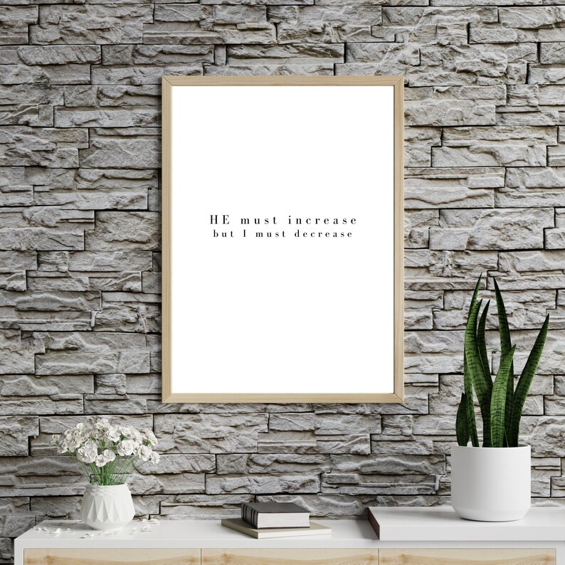 Puede incluir: Una impresi&oacute;n enmarcada con fondo blanco y texto negro que dice "HE must increase but I must decrease". La impresi&oacute;n est&aacute; colgada en una pared de piedra con una planta en maceta y un jarr&oacute;n de flores blancas en un estante de madera debajo.