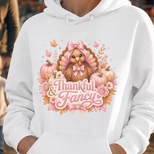 May include: A white hooded sweatshirt featuring a central graphic of a brown turkey wearing a pink bow, surrounded by pink pumpkins, autumn leaves, and flowers. The text Thankful & Fancy is printed in a pink script font below the turkey.