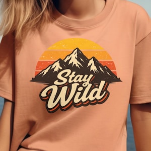 Op de afbeelding: Perzikkleurig T-shirt met een retro ontwerp met een silhouet van een bergketen tegen een zonsonderganggradiënt van oranje en geel. De woorden "Stay Wild" staan in een gestileerd lettertype onder de bergen.