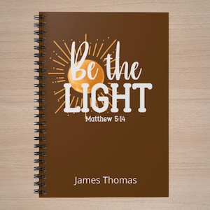 Puede incluir: Un cuaderno de espiral marrón con la inscripción blanca "Be the LIGHT" y un sol amarillo. El texto "Matthew 5:14" y el nombre "James Thomas" también son visibles en la portada.