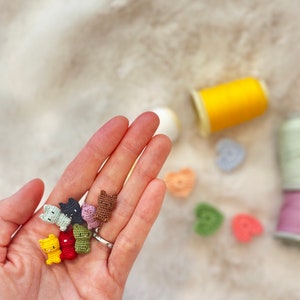 May include: A hand displaying a selection of miniature, colourful crocheted cat figures. The cats are in shades like yellow, red, green, and brown. In the background are thread spools and small crocheted hearts.