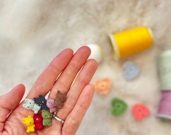 May include: A hand holding a collection of small, colorful crocheted cat figurines. The cats are in various colors, including yellow, red, green, and brown. In the background are spools of thread and small crocheted hearts.