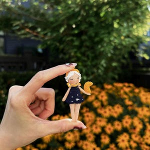 May include: A small, handmade crochet doll with a navy blue dress adorned with white star accents. The doll has a yellow scarf and a crescent moon accessory. The doll is held in front of a background of yellow flowers and green foliage.