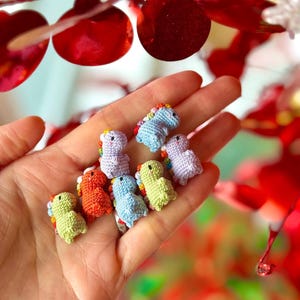 May include: A hand holds several small, crocheted dinosaur figurines in various colors, including green, orange, blue, and purple. Each dinosaur has a rainbow-colored mane. The background features red decorative elements.