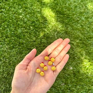 May include: A hand holds several small, yellow crocheted chicks with red crests. The chicks are evenly spaced and appear to be handmade. The background is a blurred green, suggesting grass.