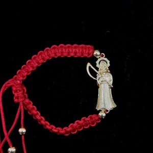 May include: A red macrame cord bracelet featuring a gold-toned metal charm of a hooded figure holding a scythe. The bracelet includes small gold-toned beads on the adjustable cord ends, creating a detailed piece of jewelry with a woven texture.