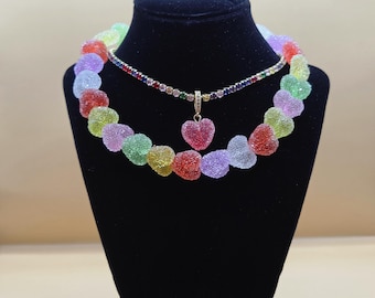 Puede incluir: Un collar con una hilera de cuentas en forma de corazón con textura de azúcar en tonos rojo, verde, amarillo y morado, combinado con un segundo collar de cadena dorada fina con piedras redondas de colores y un colgante en forma de corazón.