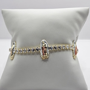 May include: A gold-toned bracelet featuring a central Virgin Mary pendant and smaller pendants on each side. The bracelet is adorned with clear, sparkling stones and rests on a white pillow. The bracelet has a delicate design.