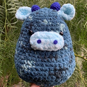 Pode incluir: Um brinquedo de pelúcia de vaca de crochê azul e branco com chifres azuis e olhos pretos. A vaca tem um nariz e uma boca azul claro.