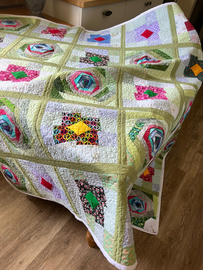 Peut inclure: Une courtepointe en patchwork vert, blanc et multicolore avec un motif floral. La courtepointe est faite de carr&eacute;s et de rectangles de tissus diff&eacute;rents, et elle a une bordure blanche.